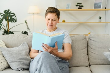 Fototapeta premium Attractive middle-aged woman with a lovely smile sitting on a sofa in the living room clutching a book.