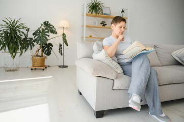 Obraz premium Middle-aged housewife sitting on sofa with book, free time retirement, hobby.