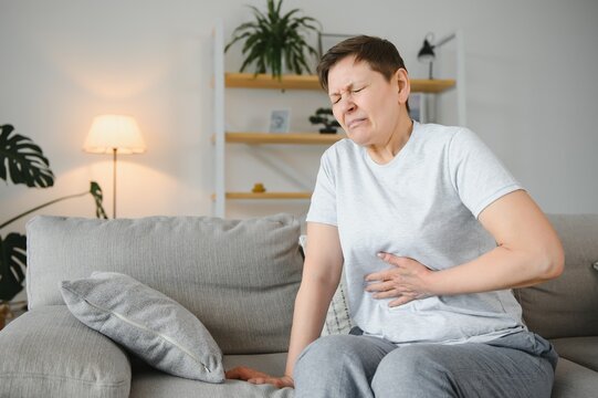 Middle-aged woman sitting on bed feels unhealthy touch stomach suffers from severe crampy abdominal pain