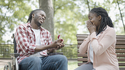 Black man in wheelchair making proposal to surprised girlfriend in park, engagement