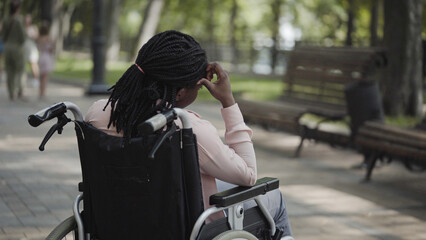 Tired African-american woman with disability sitting in wheelchair alone in park, back view