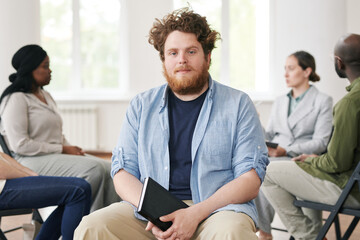 Young bearded man in casualwear holding notebook in black leather cover and looking at camera...