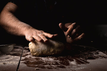 Unshaven man prepares pizza dough on the kitchen table against a dark background.