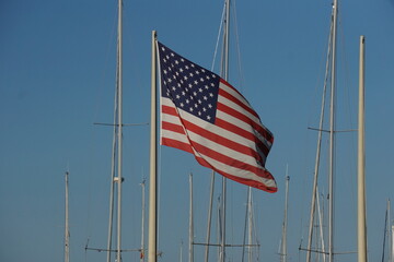Bandiera degli Stati Uniti d'America, con strisce rosse e bianche orizzontali e stelle bianche su un rettangolo blu, con gli alberi delle barche a vela e il cielo azzurro sullo sfondo