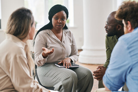 Young female psychotherapist giving advice to one of patients during session while sitting in front of group of people with problems