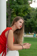 Portrait of blonde attractive young lady in elegant red dress on park background. Beautiful woman resting in park. Vertical frame.