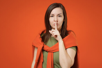 Young smiling happy woman 20s wearing khaki t-shirt tied sweater on shoulders say hush be quiet with finger on lips shhh gesture isolated on plain orange background studio. People lifestyle concept.