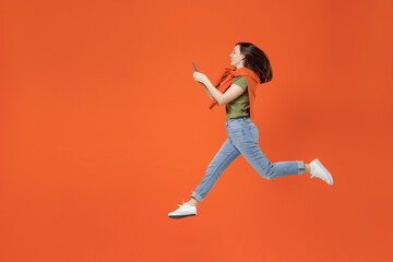 Fototapeta na wymiar Full body side view young smiling happy woman 20s in khaki t-shirt tied sweater on shoulders jump high hold in hand use mobile cell phone isolated on plain orange background. People lifestyle concept.