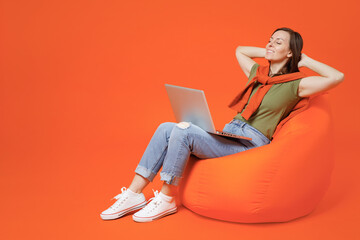 Full body young relaxed happy woman 20s wearing khaki t-shirt sweater sit in bag chair hold use work on laptop pc computer hold hands behind neck isolated on plain orange background studio portrait