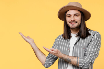 Young happy man he 20s wearing striped grey shirt white t-shirt hat pointing hands arms indicate aside on workspace area copy space mock up isolated on plain yellow color background studio portrait