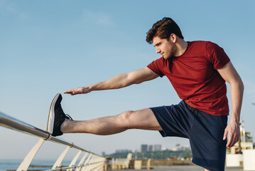 Side profile view young strong sporty fit sportsman man 20s wear sports clothes doing stretch exercise for legs warm up training at sunrise sun dawn over sea beach outdoor on pier seaside in morning.