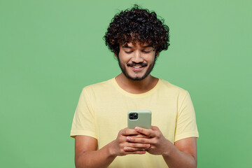 Young smiling happy Indian man 20s in basic yellow t-shirt hold in hand use mobile cell phone chatting online isolated on plain pastel light green background studio portrait. People lifestyle concept.