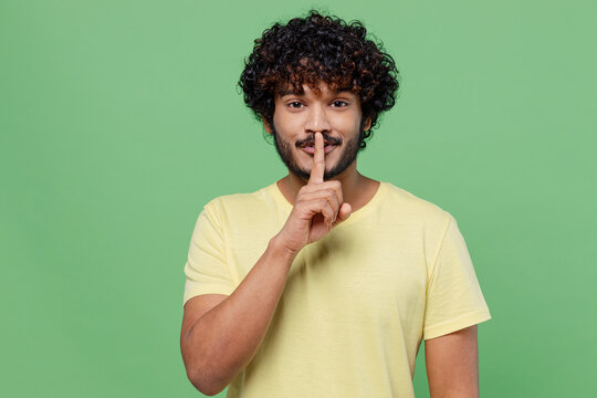 Young secret Indian man 20s wearing basic yellow t-shirt say hush be quiet with finger on lips shhh gesture isolated on plain pastel light green background studio portrait. People lifestyle concept.