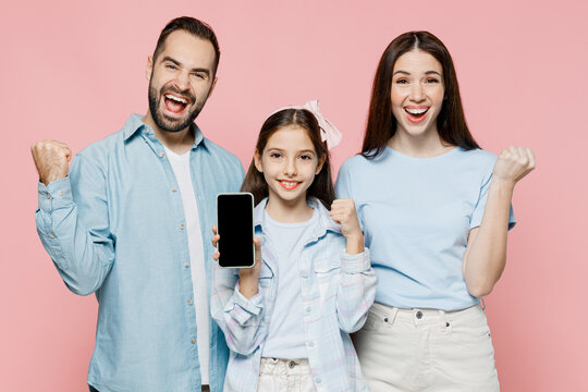 Young parents mom dad with child kid daughter teen girl in blue clothes hold use mobile cell phone screen workspace area do winner gesture isolated on plain pastel pink background. Family day concept.
