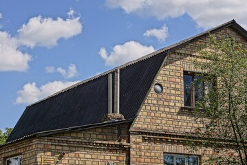 Fototapeta premium two gray chimneys on the slate roof of a brown brick private house with windows against a blue sky
