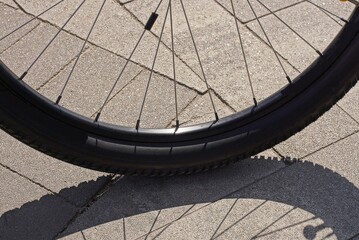 part of a black bicycle wheel with metal spokes stands on a gray pavement in the street