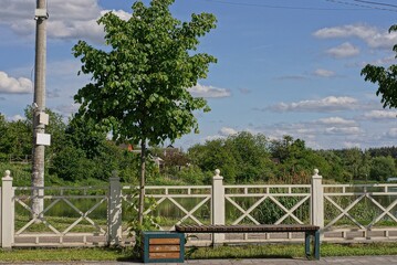 one brown empty wooden bench stands on a alley with green grass by a white fence in a park against the background of trees and blue sky