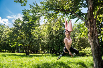 Happy physically developed woman practices yoga on aerial canvases on a tree.