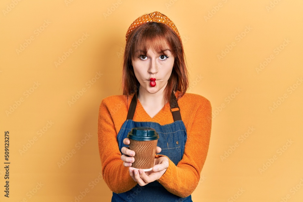 Poster Redhead young woman wearing waitress apron holding take away cup of coffee making fish face with mouth and squinting eyes, crazy and comical.