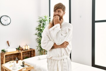 Redhead man with long beard wearing bathrobe at wellness spa looking stressed and nervous with hands on mouth biting nails. anxiety problem.