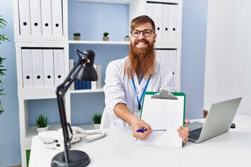 Obraz premium Young redhead man wearing doctor uniform holding clipboard at clinic