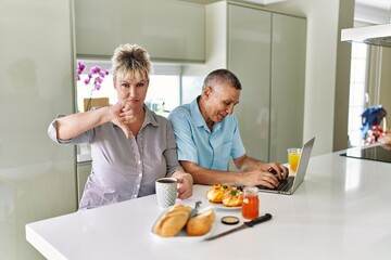 Middle age caucasian couple eating breakfast at home using laptop with angry face, negative sign showing dislike with thumbs down, rejection concept