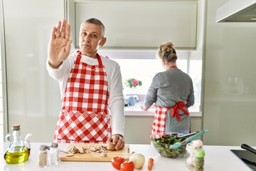 Middle age caucasian couple cooking healthy salad with open hand doing stop sign with serious and...