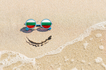Fototapeta na wymiar A painted smile on the beach and sunglasses with the flag of the Bulgaria. The concept of a positive holiday in the resort of the Bulgaria.