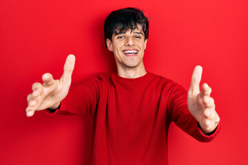 Handsome hipster young man wearing red winter sweater looking at the camera smiling with open arms for hug. cheerful expression embracing happiness.