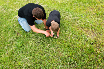 Happy middle aged father playing with his little son in the park. Son holds a magnifying glass and examines the beetle.