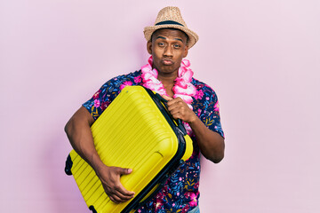 Young black man wearing summer hat holding cabin bag looking at the camera blowing a kiss being...