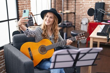 Young blonde woman musician making selfie by smartphone holding classical guitar at music studio