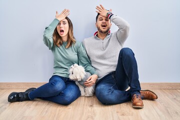 Young hispanic couple sitting on the floor with dog surprised with hand on head for mistake,...