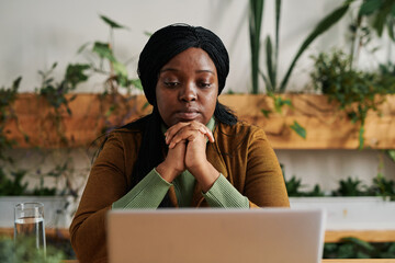 Young serious female psychotherapist sitting in front of laptop screen in office and listening to...