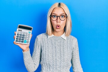 Beautiful blonde woman showing calculator device scared and amazed with open mouth for surprise,...