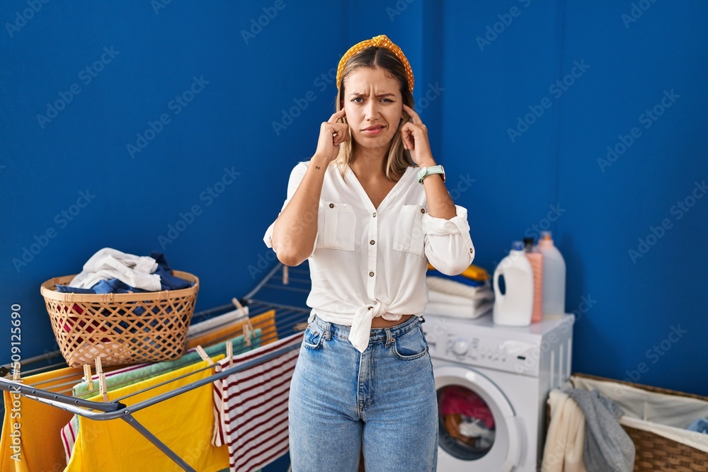 Sticker Young blonde woman at laundry room covering ears with fingers with annoyed expression for the noise of loud music. deaf concept.