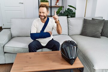 Middle age man with beard wearing arm on sling because motorbike accident surprised with an idea or question pointing finger with happy face, number one