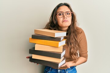 Young hispanic girl holding a pile of books clueless and confused expression. doubt concept.