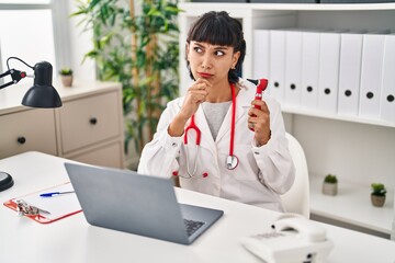 Young hispanic doctor woman holding ear thermometer serious face thinking about question with hand on chin, thoughtful about confusing idea