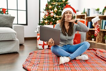 Obraz na płótnie Canvas Young latin woman using laptop sitting by christmas tree cutting throat with hand as knife, threaten aggression with furious violence