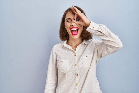 Young beautiful woman standing casual over blue background doing ok gesture with hand smiling, eye looking through fingers with happy face.