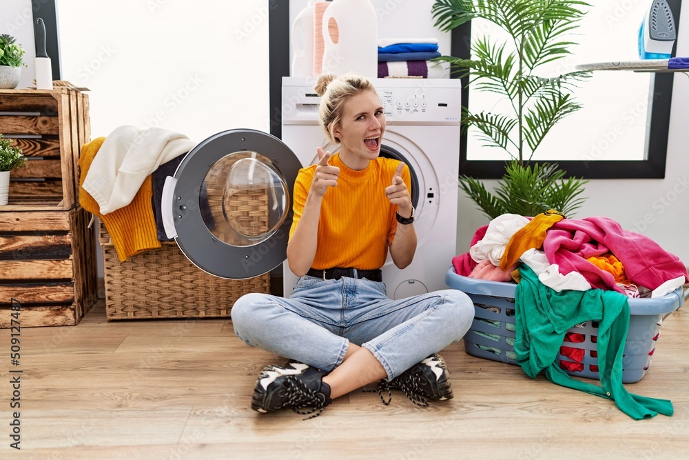 Poster Young blonde woman doing laundry sitting by washing machine pointing fingers to camera with happy and funny face. good energy and vibes.