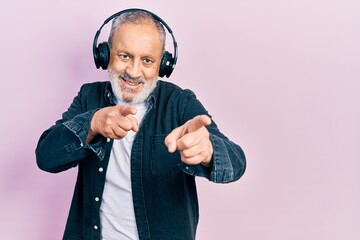 Handsome senior man with beard listening to music using headphones pointing fingers to camera with happy and funny face. good energy and vibes.