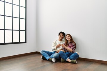 Young hispanic couple toasting with champagne sitting on the floor at empty new home.