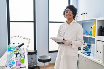 Naklejka na ściany i meble Young hispanic man wearing scientist uniform writing on clipboard at laboratory