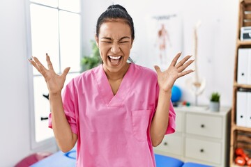 Young hispanic woman working at pain recovery clinic celebrating mad and crazy for success with...