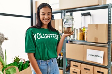 Young hispanic volunteer woman holding charity jar with money looking positive and happy standing and smiling with a confident smile showing teeth