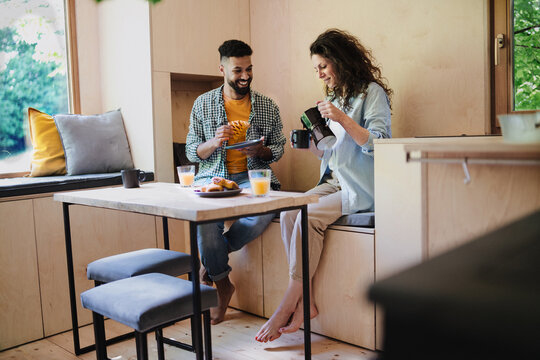 Happy couple having breakfast, sitting, resting and talking indoors in a tree house, morning routine. - Powered by Adobe