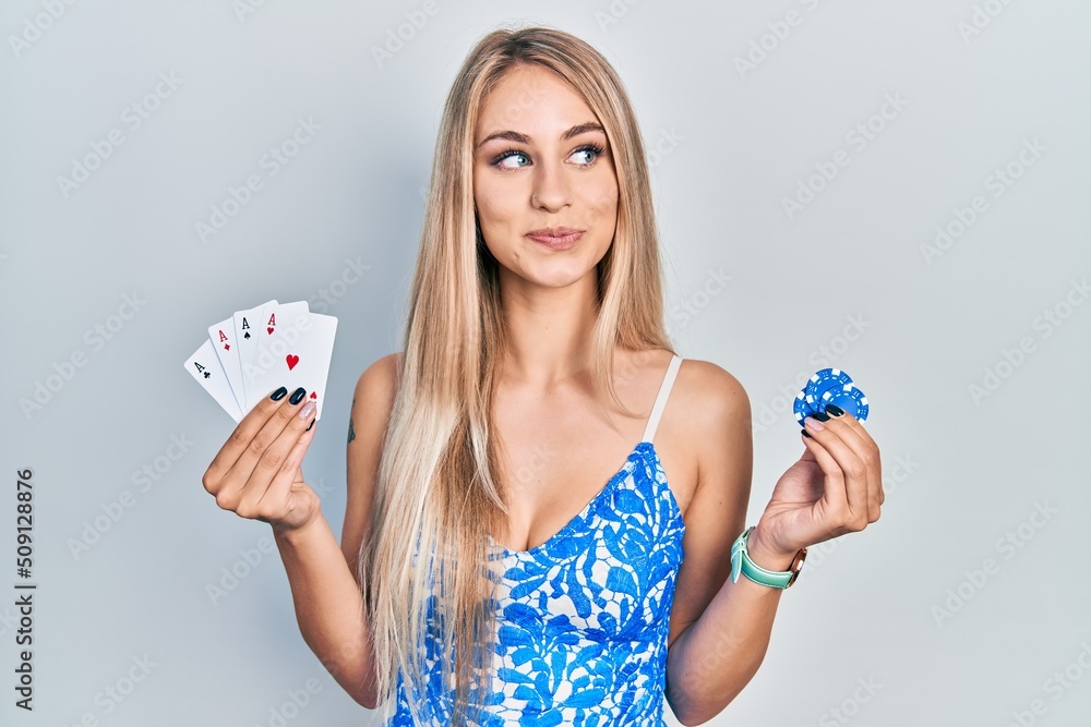 Wall mural Young beautiful caucasian woman holding poker cards and chips smiling looking to the side and staring away thinking.