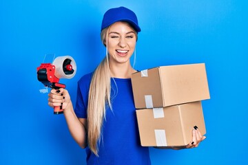 Young caucasian woman holding packages and packing tape winking looking at the camera with sexy...